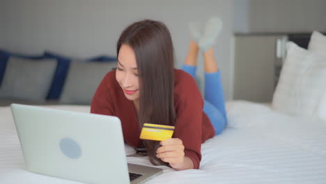 asian woman type in card information in online shopping mall using laptop computer while laying on a comfortable bed, slow motion close-up