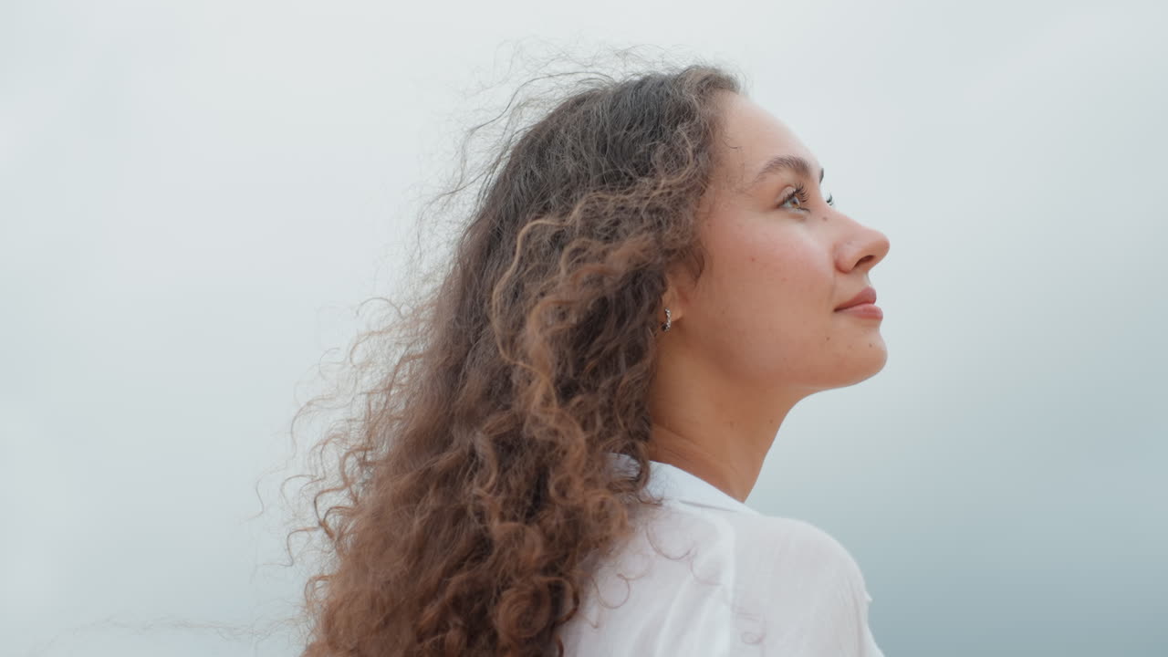 Woman with flowing hair views serene coast. Back view of lady watching peaceful island scene by sea. Profile of woman with windswept hair gazing across calm ocean and rugged cliffs