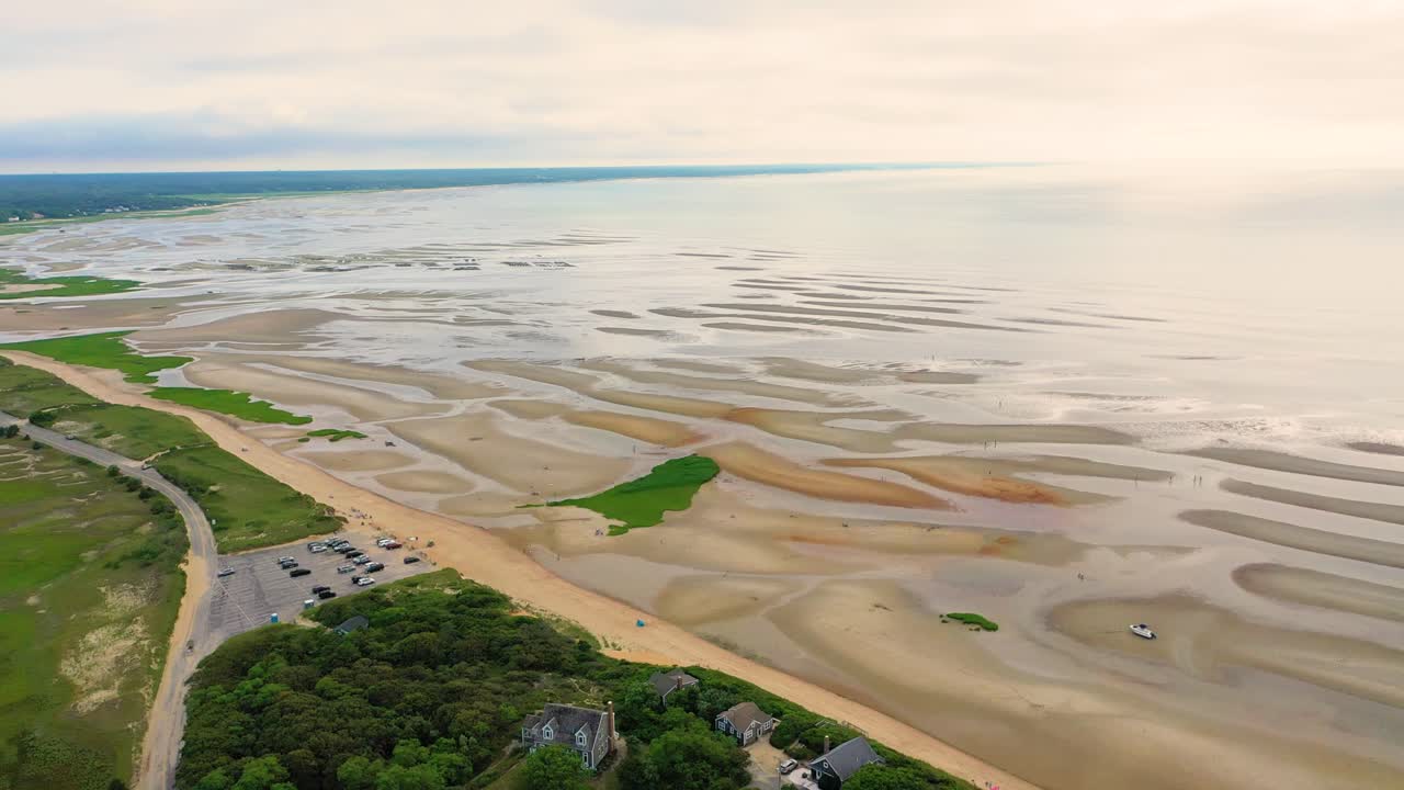 Drone footage captures expansive tidal flats at low tide with rippled sand patterns, reflective tide pools, and green marsh patches, while houses and a parking lot line the public beach access