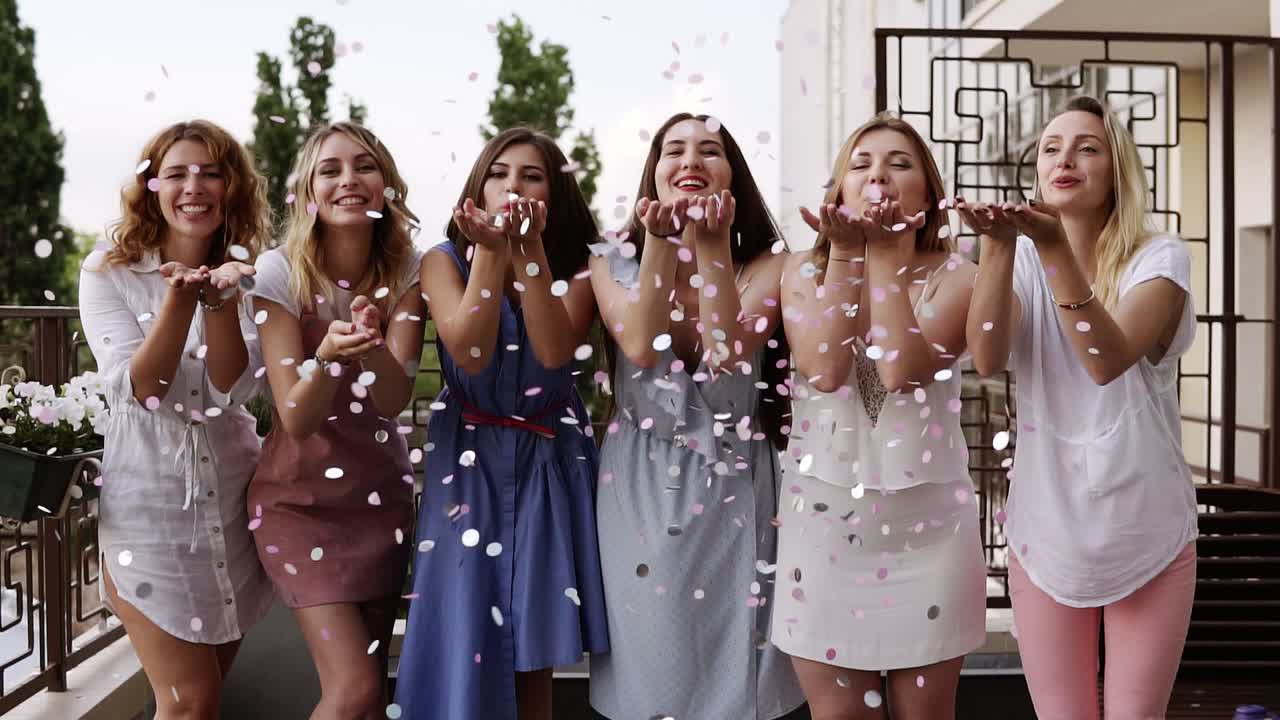 Front view of six attractive caucasian young women playfully blowing bright colored confetti from their hands together. stylish clothes. Bachelorette party. slow motion