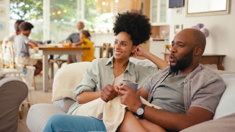 Pareja-En-Casa-En-Un-Sofá-Transmitiendo-Un-Programa-O-Una-Película-A-La-Televisión-Con-Una-Familia-Multigeneracional-En-Segundo-Plano