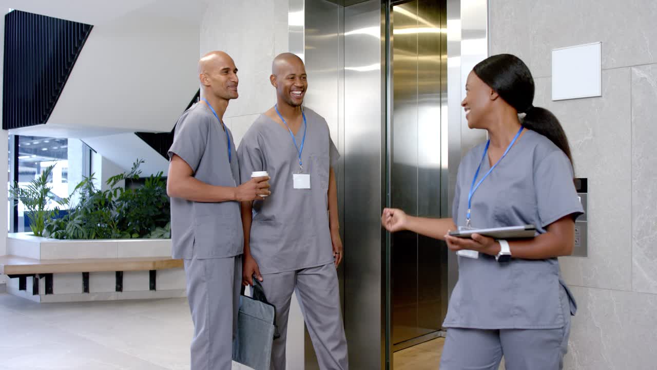 Profissional de saúde afro-americana em uniforme cinza apertando botão de chamada e entrando no elevador com tablet e bolsa.