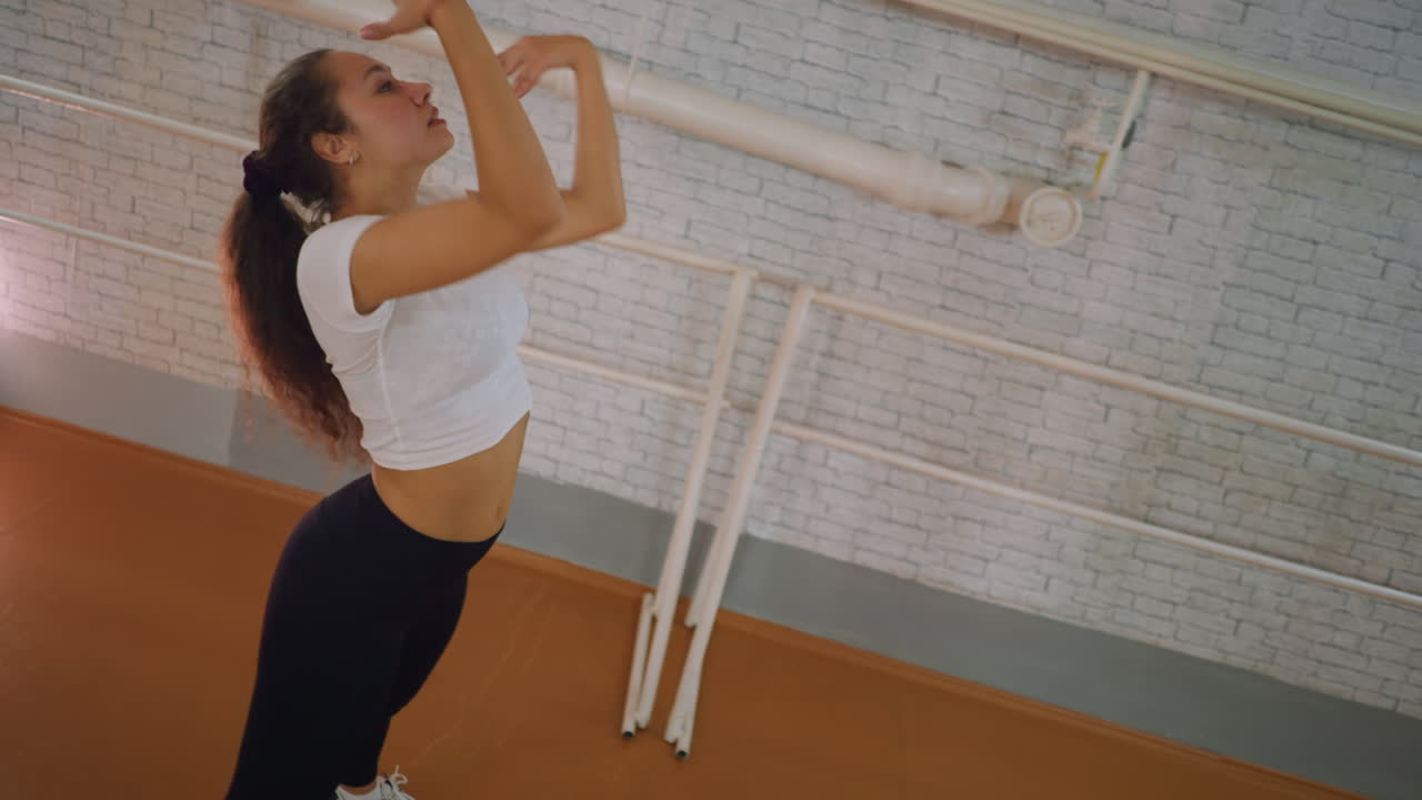Side view of young woman in white top and black leggings dancing in studio placing hand on knee twisting body flipping hair with energy expressive motion rhythm confidence and athletic performance