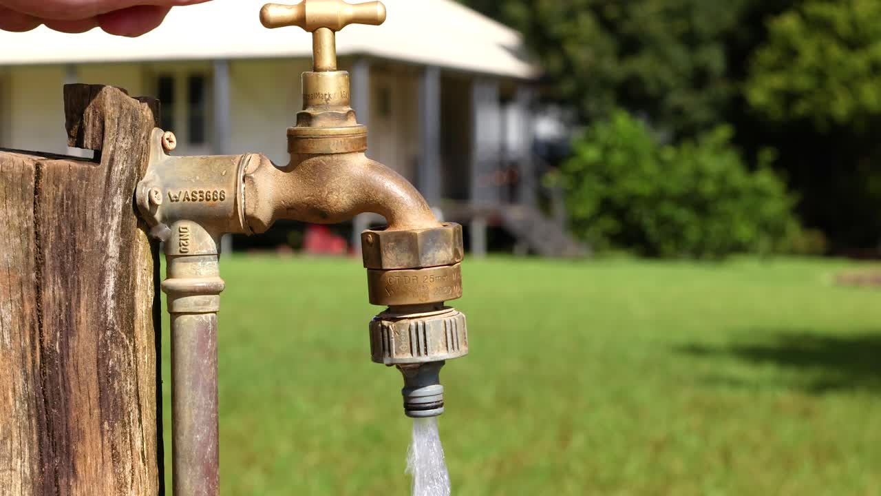 A hand turns a brass faucet on a wooden post, releasing water in a sunny farm setting