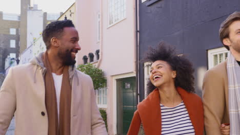 Two-multi-cultural-couples-arm-in-arm-as-they-walk-along-cobbled-mews-street-on-visit-to-city-in-autumn-or-winter-carrying-sale-shopping-bags---shot-in-slow-motion