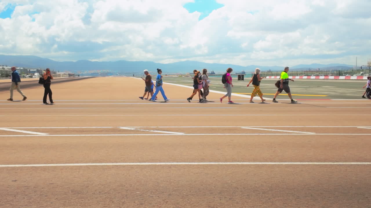 People walking across Gibraltar's airport runway, an unusual and unique urban feature, under a bright blue sky with mountains in the background