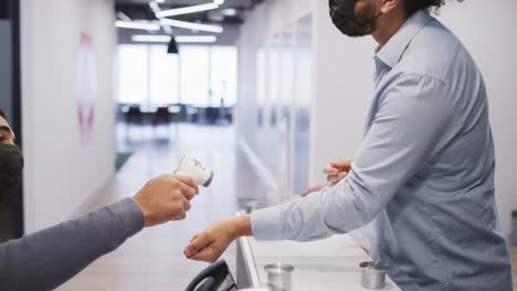 Mixed-race-man-wearing-face-mask-has-temperature-checked-by-male-colleague-at-office-reception-desk