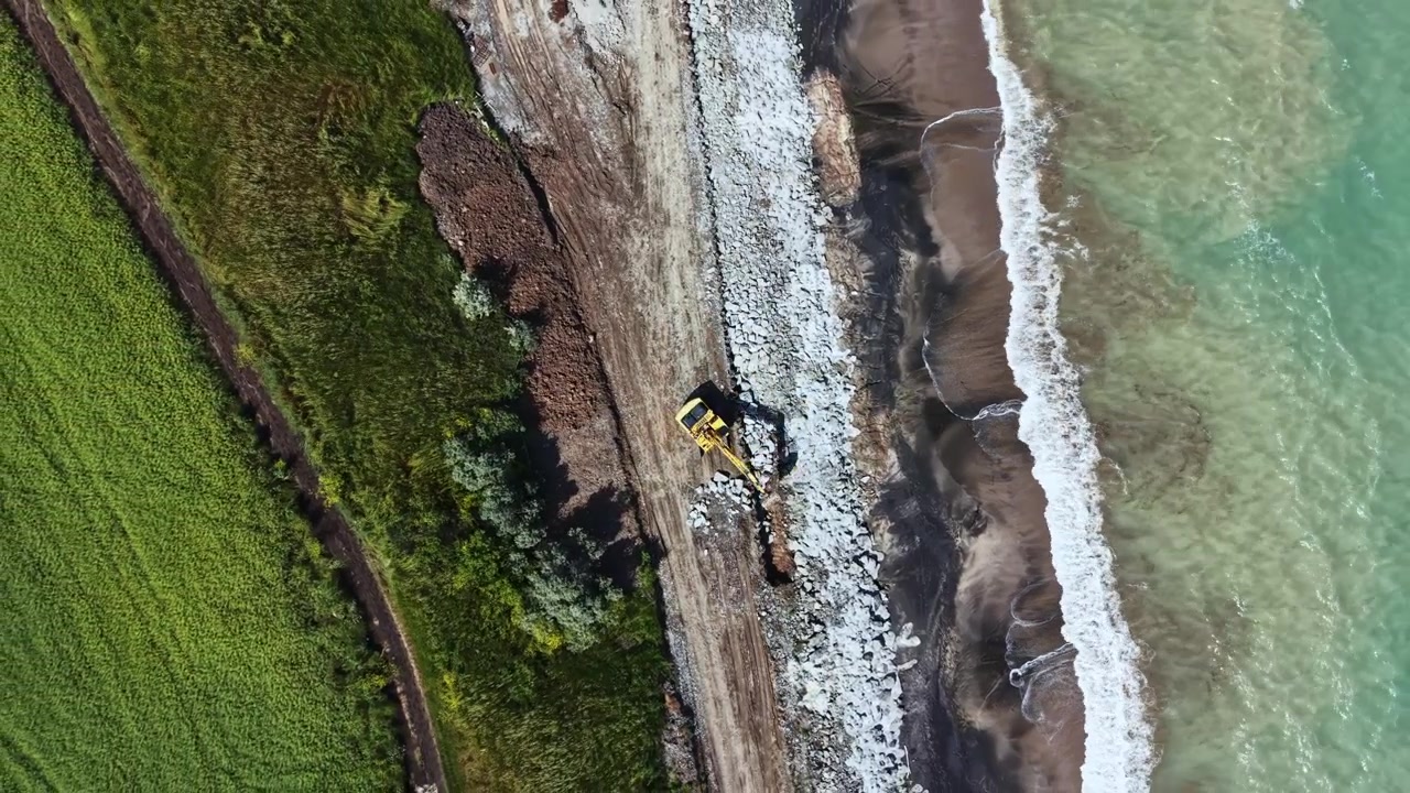 Máquinas pesadas trabalham na orla para restaurar a área da praia perto de um corpo d'água durante o dia, com ondas visíveis e vegetação na paisagem.