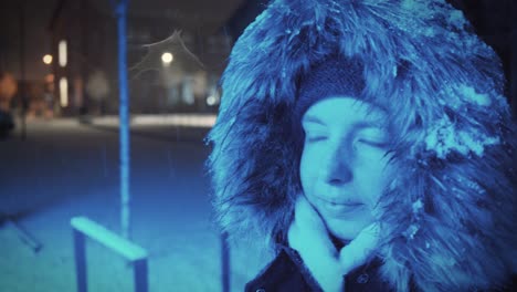 young woman with a snow-covered fur hood is standing in a blue light in a wintry landscape at night