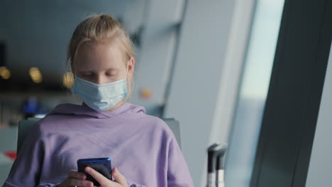 a child in a protective mask sits in the airport waiting room, using a smartphone. traveling during the coronavirus pandemic