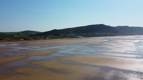 descending drone shot on rhossili bay with grassy sand dunes and moorland fields backdrop with hazy blue sky 4k