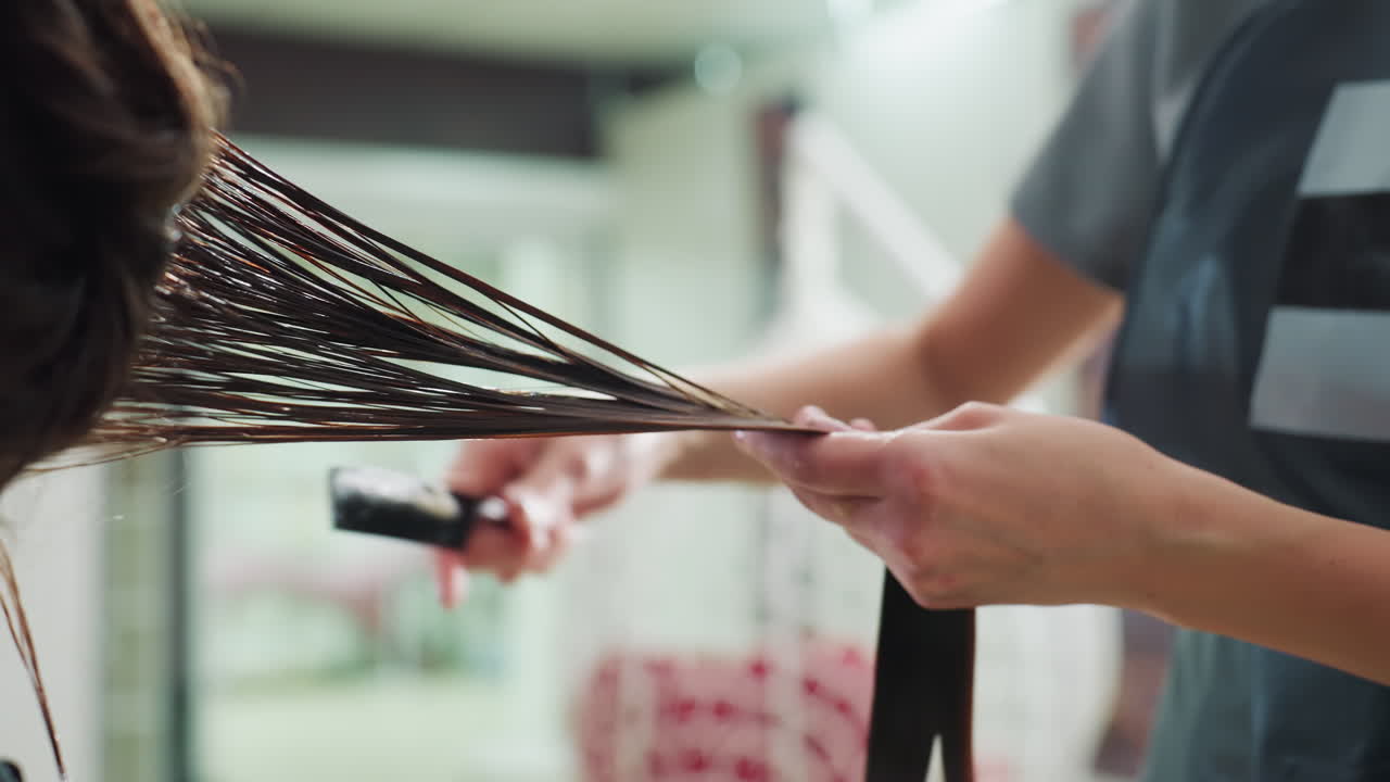 클로즈업 스타일리스트가 젖은 긴 머리의 끝을 다듬고 빗질하는 모습이에요. 손은 빗과 가위로 머리를 잡고 있고, 밝은 살롱 선반이 배경이에요. 깔끔한 마무리와 건강한 머리끝에 초점을 맞춘 섬세한 기술이 돋보여요.