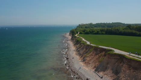 Luftaufnahme-Der-Deutschen-Küste-Mit-Blick-Auf-Die-Ostsee,-Weitläufiges-Grünes-Feld-An-Der-Küste-Mit-Vorbeigehenden-Passanten,-Klarer-Himmelstag,-Vorwärts