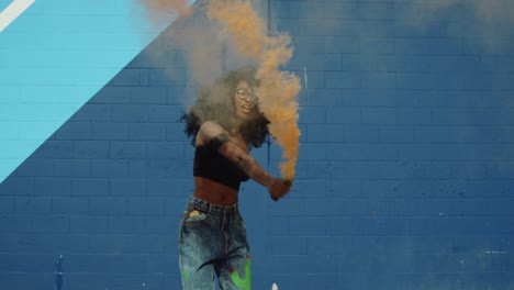 beautiful young woman holding colorful smoke grenade dancing outside against blue wall