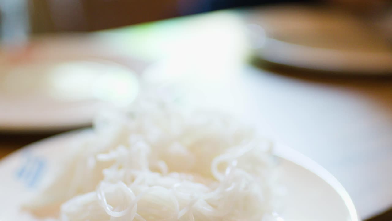 Hand lifts soft Thai rice noodles from plate, shallow depth of field, warm ambient lighting