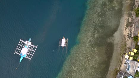 Bangka-boat-drifting-away-from-island-in-Philippines,-overhead-drone-shot-with-coral-reef-line