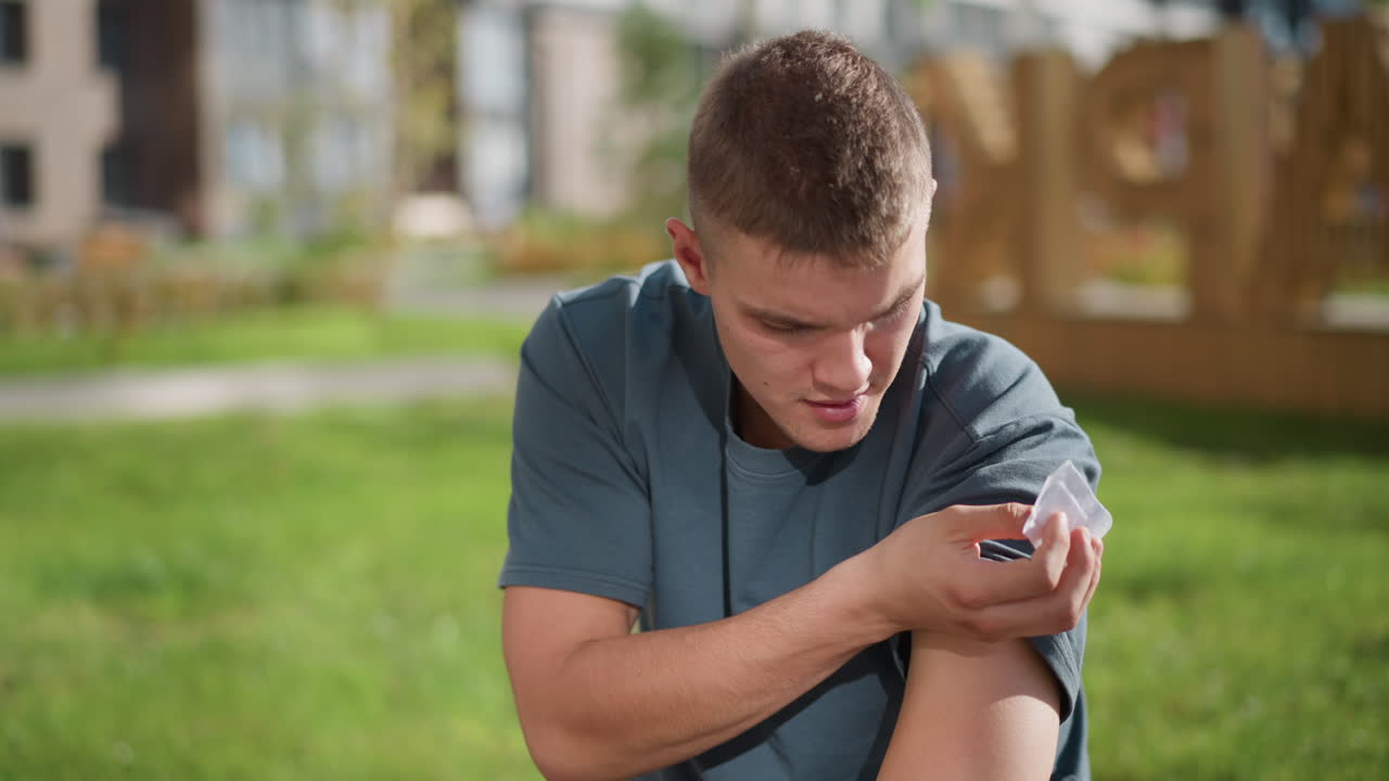 young boy seated in sunlight lifts shirt sleeve and carefully applies nicotine patch to upper arm while surrounded by soft blur of green outdoor environment suggesting self-care and wellness action