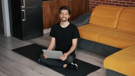 portrait of a man sitting on the mat on the floor with laptop at home