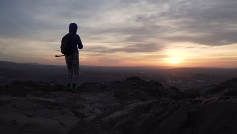 Static-shot-of-girl-walking-to-the-edge-of-a-mountain-to-take-a-photo-with-smartphone-during-beautiful-sunset-of-Edinburgh-cityscape-with-rocky-cliffs-in-foreground