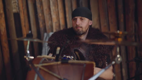 bearded man with fur plays drums sitting near rough logs