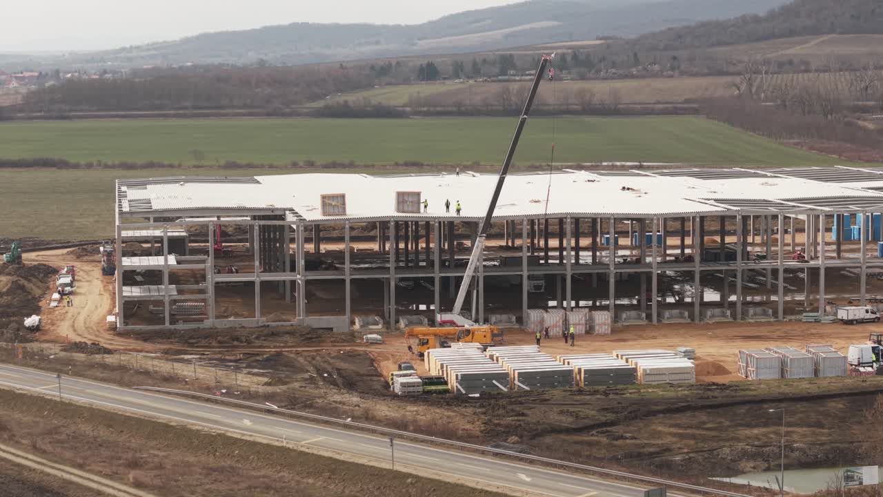 Un magazzino industriale in costruzione in un'area rurale, caratterizzato da una struttura in cemento e installazione di un tetto metallico. Una gru sta sollevando materiali mentre gli operai edili sono sul tetto