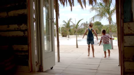 happy caucasian siblings running and raising hands at swimming pool at beach house with palm trees