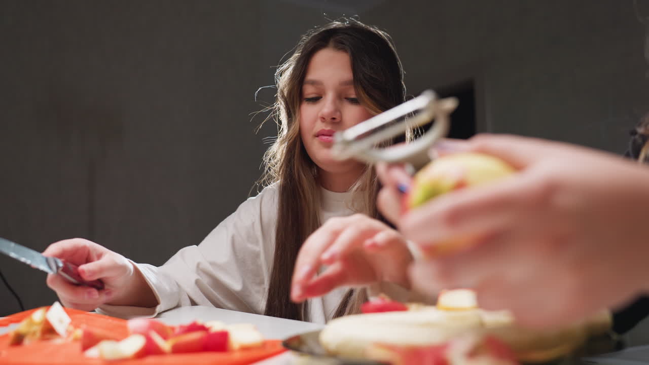 Girlfriends in kitchen talk while one chops apples on orange cutting board and other peels apple skin with peeler; teen students prepare healthy snack under bright light, close foreground hands
