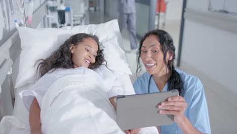 Diverse-female-doctor-and-child-patient-using-tablet-in-corridor-at-hospital,-in-slow-motion