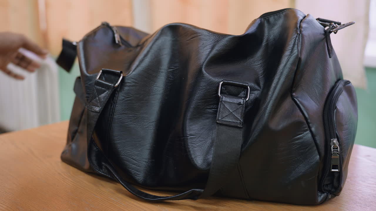 Close up of black bag being dropped on wooden table while strap is adjusted by person in sportswear, capturing preparation moment and detail of equipment handling in indoor environment