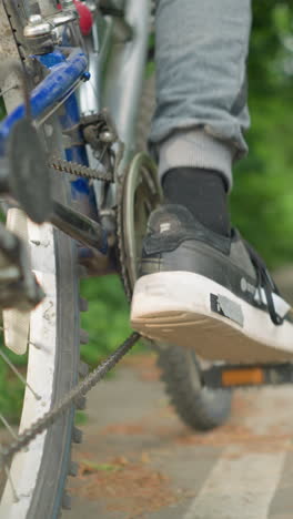 close-up back view of someone tapping the pedal of the bicycle backwards with their foot on a paved path, the blurred background features lush greenery and road markings