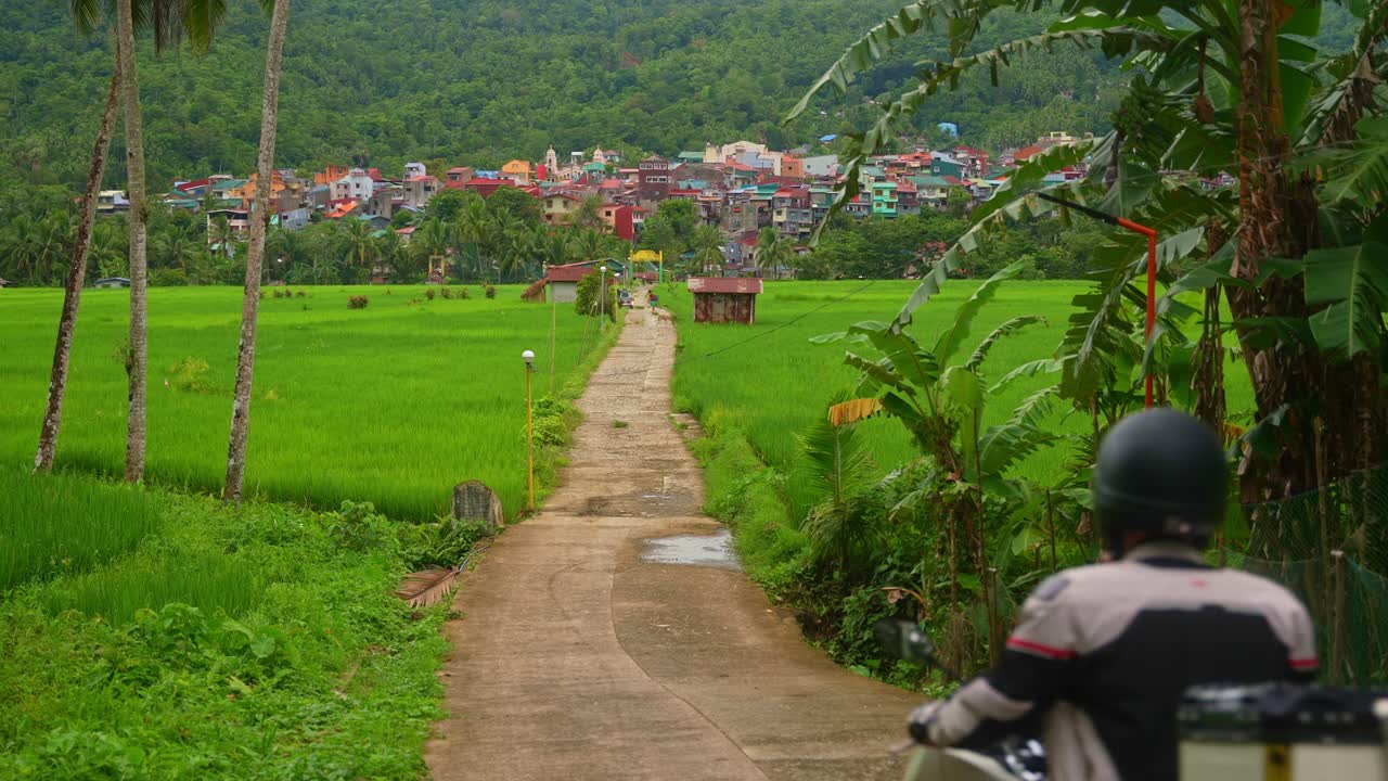 A shot of motorcycle rider wearing helmet drives into frame along Mahabang Tanaw through lush rice fields, heading toward colorful village houses in Quezon Province Philippines