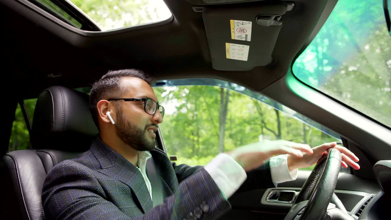 Side view shot of young Middle Eastern man in suit and eyeglasses driving car down city road singing and dancing to music, wireless headphones in his ears