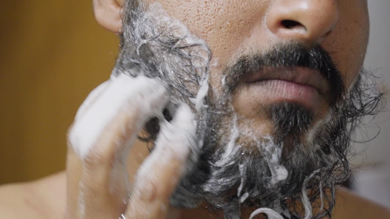 A close-up of a man with a beard gently washing it with soap, focusing on his hands and beard as the lather creates a clean, soft texture