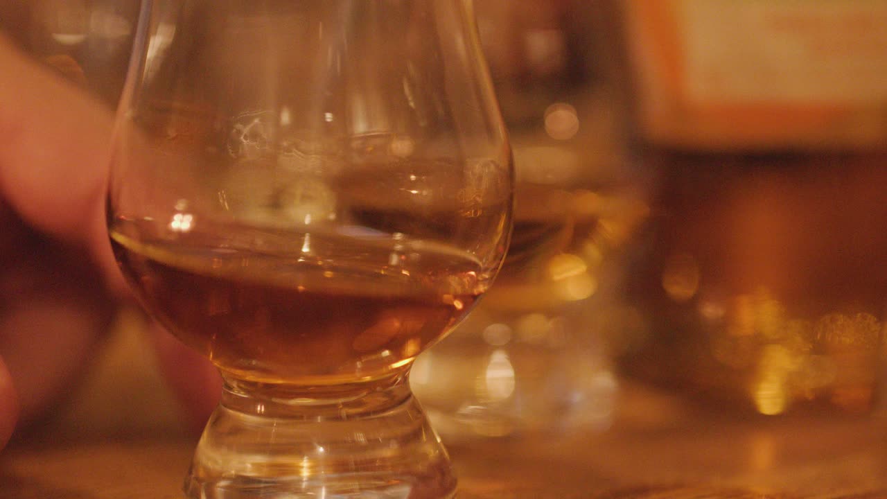 A hand gently swirls a whisky glass on a wooden table, surrounded by bottles, under soft, warm lighting with shallow depth of field