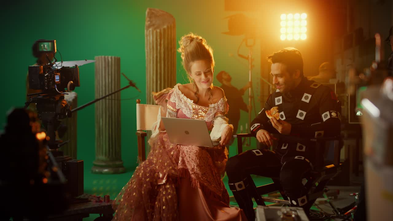 르네상스 드레스를 입은 아름다운 미소 짓는 여배우와 모션 처 슈트를 입은 배우가 점심 휴식을 취하고 의자에 앉아 노트북을 사용하고 이야기합니다.