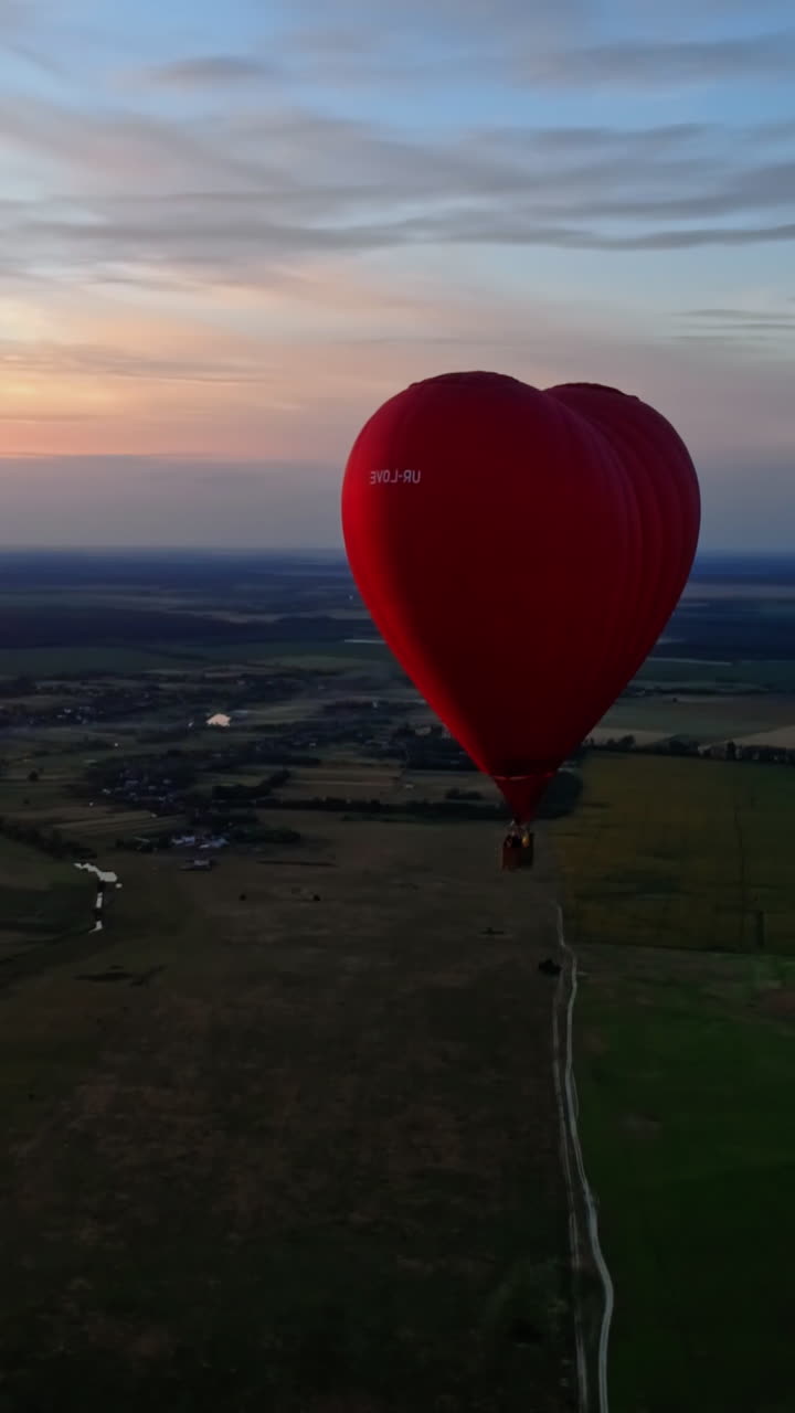 Red aerostat against the setting sun. Hot air balloon flying in the sky over the field in the countryside. Hot air balloon in the form of a heart. Aerial view. Vertical video