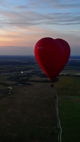 red aerostat against the setting sun. hot air balloon flying in the sky over the field in the countryside. hot air balloon in the form of a heart. aerial view. vertical video