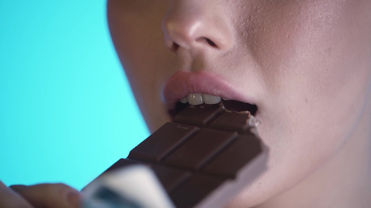 Close-Up Of Woman's Mouth Eating Chocolate Bar Against Blue Background, Studio Shot