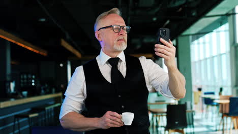 bearded man having coffee break and using his phone. positive man taking selfie or making video.