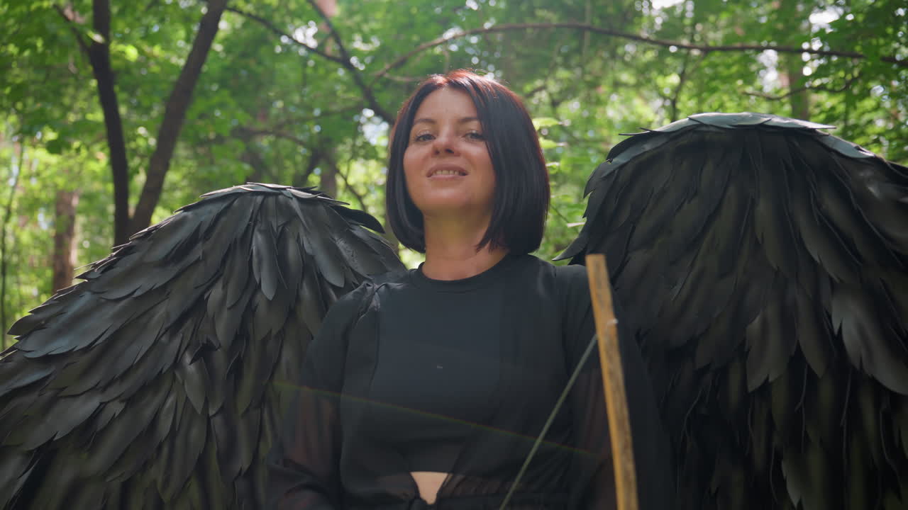 Forest warrior with majestic black wings stands holding bow under green canopy, sunlight highlighting her calm yet powerful gaze, mysterious fantasy scene blending strength and ethereal beauty