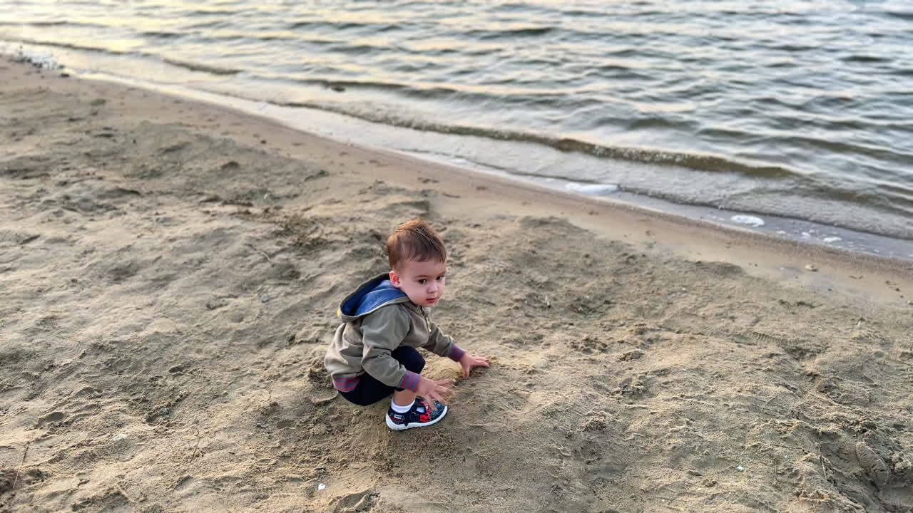 Baby boy in shorts and hoodie plays with sand on the shore. Happy kid spending time at the beach. Top view.