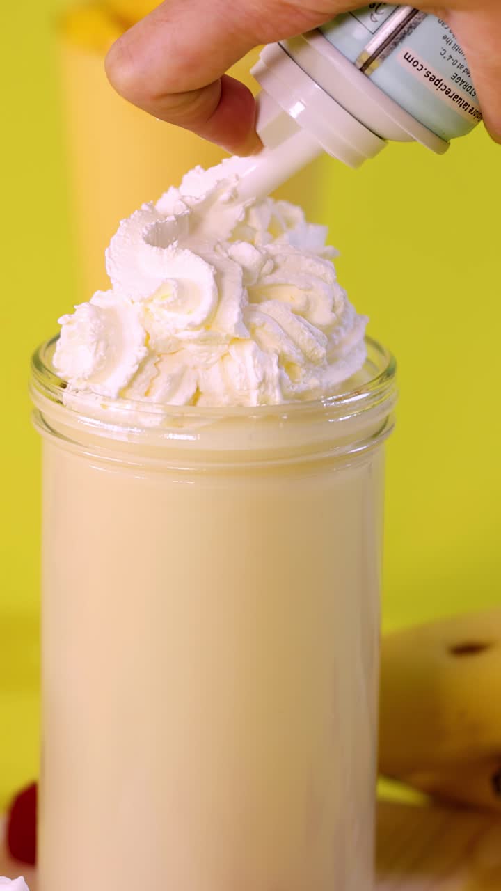 Una mano guarnisce un cremoso milkshake alla banana con panna montata, sfondo giallo brillante, primo piano, illuminazione naturale.