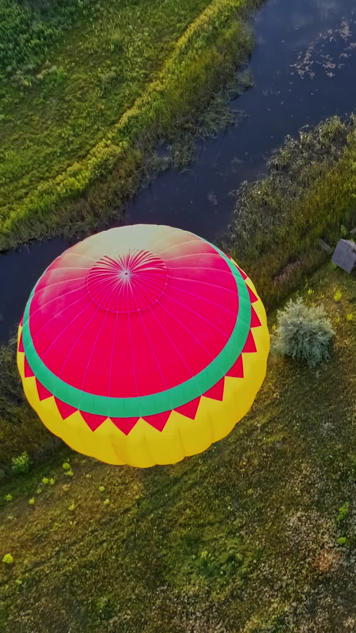 Aerostat on nature background. Colorful hot air balloon flying over the field near the small river. Outdoor activity. Top view. Camera moves down. Vertical video