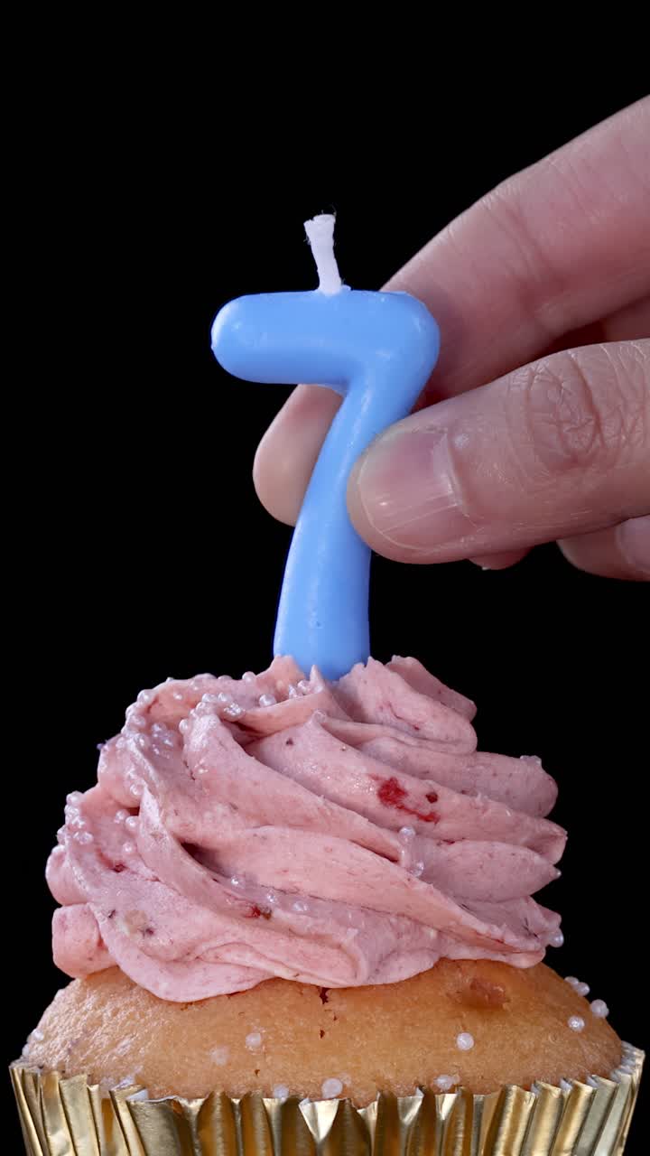 Una mano inserisce una candela di compleanno blu con il numero sette in un cupcake con glassa rosa alla fragola, sotto una luminosa illuminazione da studio e uno sfondo nero.