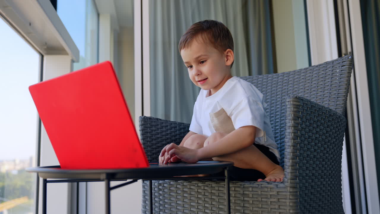 Child enjoys playing on laptop outside. A young boy sits on a balcony, reaching out joyfully toward his bright red laptop, immersed in fun and learning