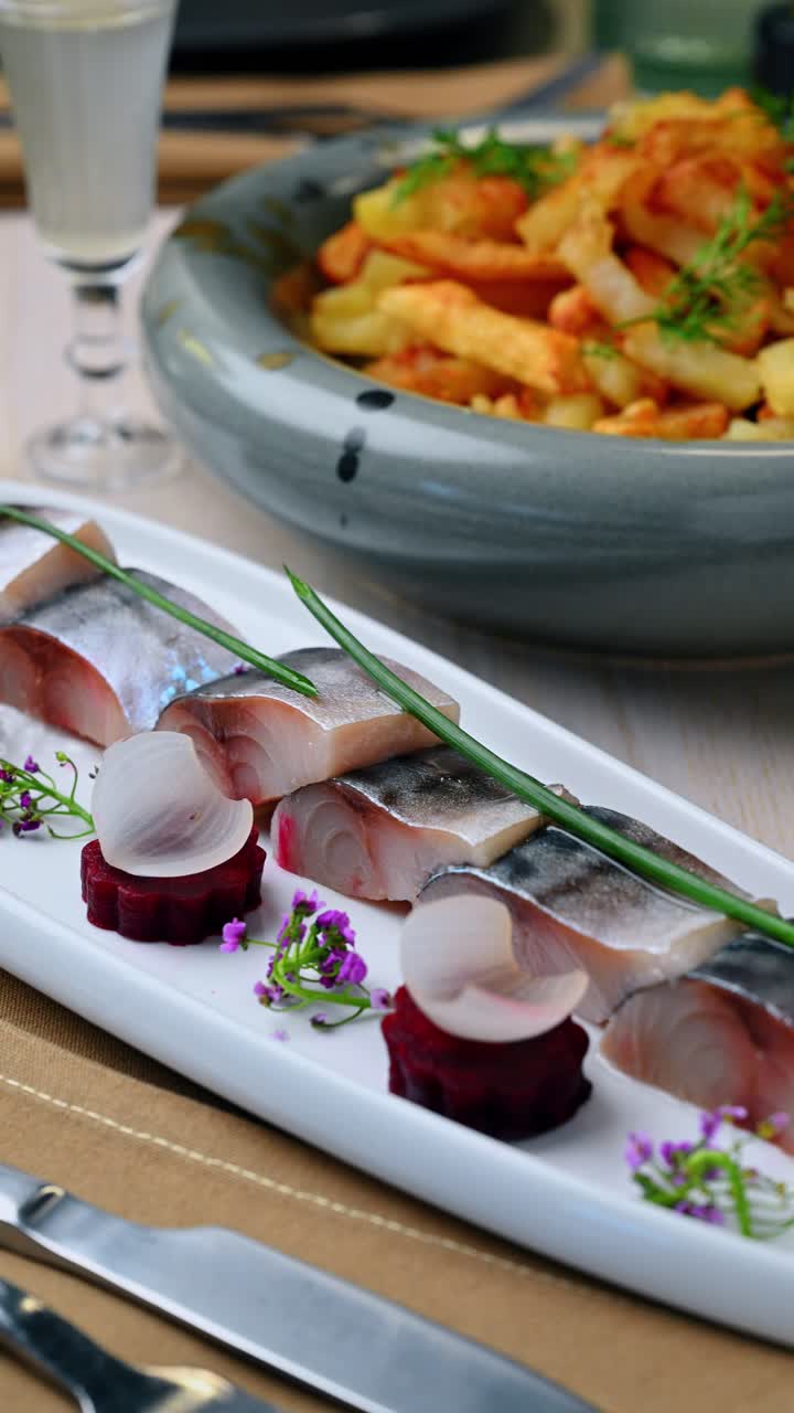 Primo piano di filetto di aringa su piatto bianco rettangolare. Vista dettagliata di fette di pesce decorate con cipolla e fiori, con ciotola di patate sullo sfondo.