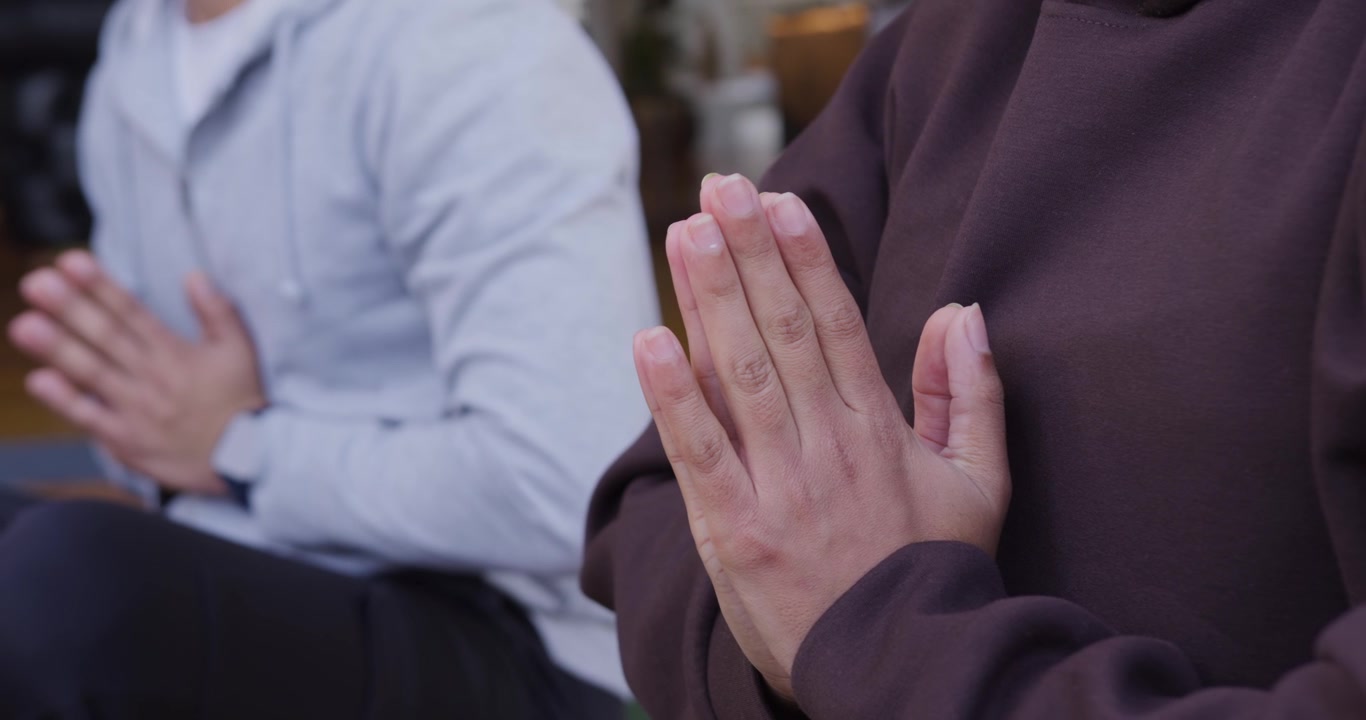 Due Persone che Meditano a Casa con le Mani Giunte, mentre la Telecamera si Allontana Rivelando Orologio e Felpe con Cappuccio