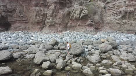 trucking-following-aerial-of-a-man-with-a-yellow-backpack-walking-along-the-seaside-traversing-boulders-and-ocean-rocks-adventuring-with-a-dramatic-cliff-in-the-background