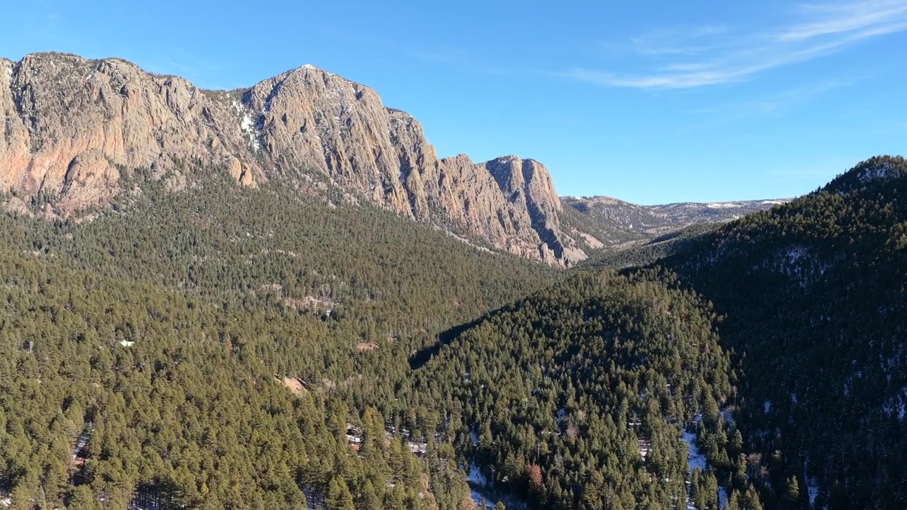 Riprese drone avanzano sopra una valle montana coperta di pini verso imponenti pareti rocciose, rivelando le Brazos Cliffs che si ergono ripidamente sopra una fitta foresta con leggere chiazze di neve e un terreno alpino accidentato