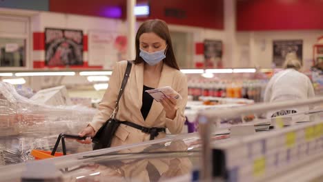 A young woman in a medical mask buys food in a supermarket during the Covid-19 coronavirus epidemic. The woman stores food during quarantine and self-isolation. Social video
